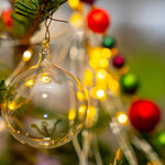 Eine durchsichtige Glaskugel, die an einem Zweig hängt. darin spiegelt sich ein weiterer Zweig und die Beleuchtung des Weihnachtsbaums. Dahinter bunte, etwas kleinere Kugeln und Beleuchtung.