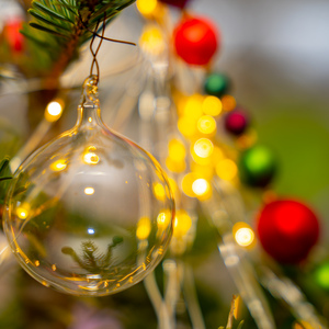 Eine durchsichtige Glaskugel, die an einem Zweig hängt. darin spiegelt sich ein weiterer Zweig und die Beleuchtung des Weihnachtsbaums. Dahinter bunte, etwas kleinere Kugeln und Beleuchtung.